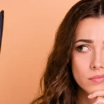 Žena promatra peglu za kosu uz zabrinut izraz lica na bež pozadini.